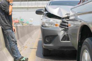 Man standing next to a car with front-end damage after a rear-end collision.